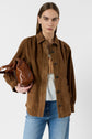 A woman with straight brown hair wears a brown suede jacket over a white top and blue jeans, holding a brown leather bag under her arm, standing against a plain white background.