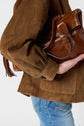 A person wearing a brown suede jacket and blue jeans holds a brown leather handbag tucked under their arm.