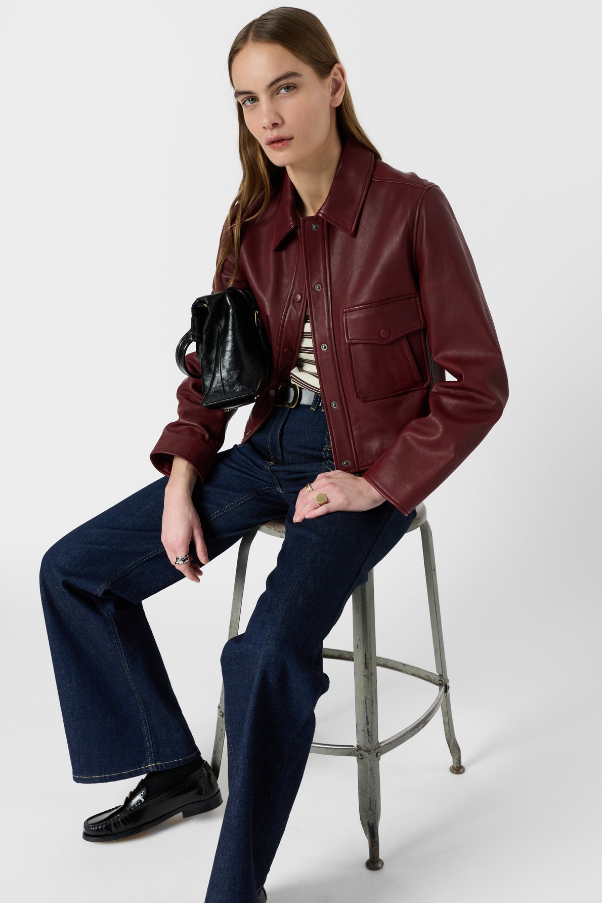 Woman in a burgundy leather jacket, striped shirt, and dark flared jeans sits on a metal stool, holding a black purse, against a plain white background.