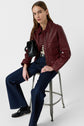 Woman in a burgundy leather jacket, striped shirt, and dark flared jeans sits on a metal stool, holding a black purse, against a plain white background.