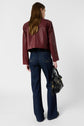 A person with long brown hair, wearing a maroon leather jacket and dark flared jeans, stands facing away and holds a black handbag in their left hand.