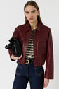 A person wearing a burgundy leather jacket, striped top, and dark jeans holds a black handbag against a plain white background.