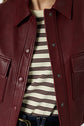 Close-up of a person wearing a burgundy leather jacket over a cream and brown striped shirt, with part of dark denim jeans and a gold belt buckle visible at the bottom.