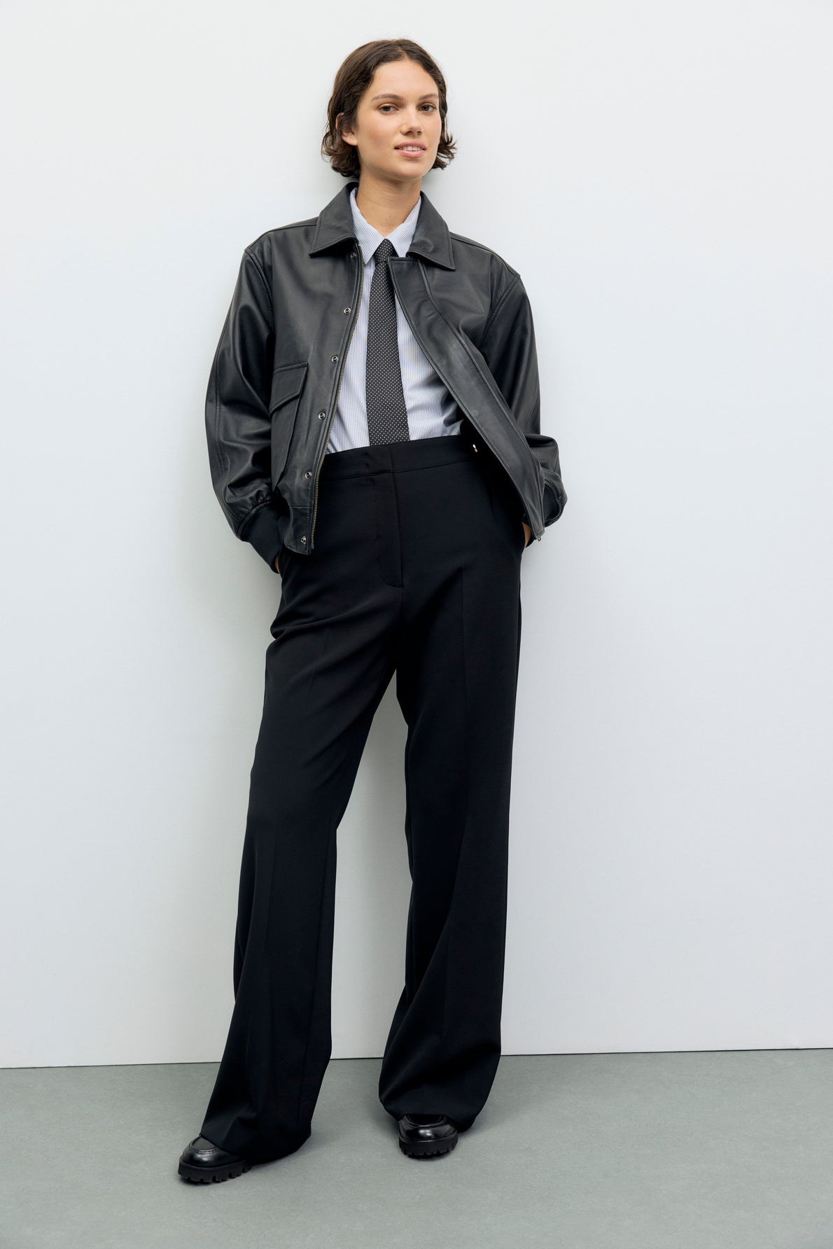 A person stands against a plain wall, wearing a black veste en cuir d’agneau, white shirt, black tie, black wide-leg pants, and black shoes. They have short, wavy hair and a relaxed, confident posture.