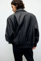 Person with short brown hair wearing a veste en cuir noire and black pants, standing with their back to the camera against a plain light background.