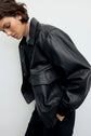 A person with short brown hair wears a veste en cuir noire over a white top, looking downward with hands in pockets, standing against a plain light background.