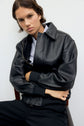 Person with short brown hair wearing a veste en cuir noir over a collared shirt, sitting with arms crossed, looking at the camera against a plain white background.