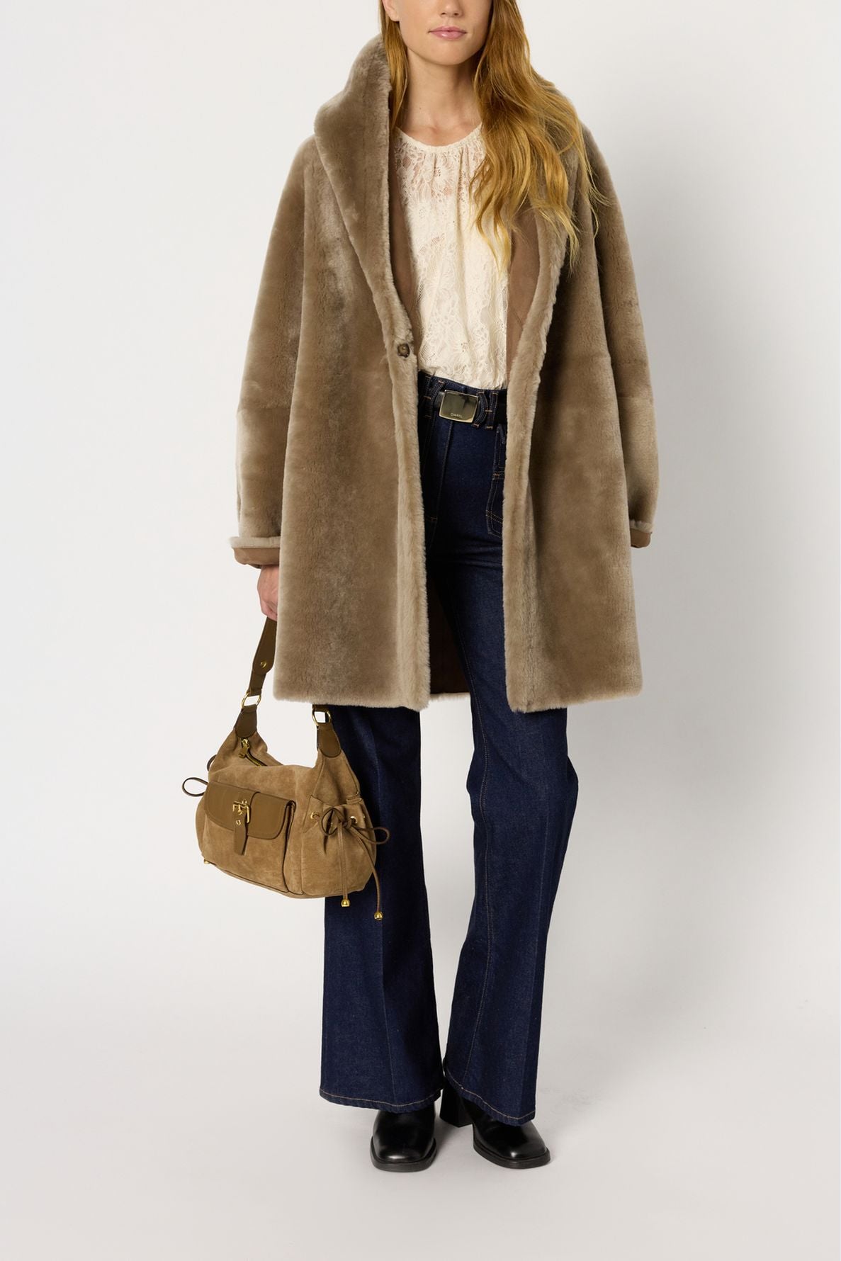 Woman wearing a tan manteau peau lainée, cream lace top, blue flared jeans, black shoes, and holding a tan handbag, standing against a plain white background.