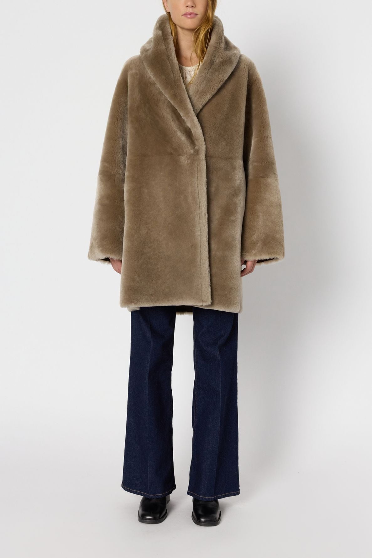 Person wearing a light brown manteau réversible in faux fur, dark blue wide-leg jeans, and black shoes, standing against a plain white background.