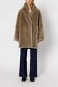 Person wearing a light brown manteau réversible in faux fur, dark blue wide-leg jeans, and black shoes, standing against a plain white background.