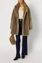 A woman wearing a tan manteau col châle faux fur coat, lace top, flared blue jeans, and black boots holds a tan suede handbag. She has long, wavy hair and stands against a plain white background.