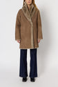Person wearing a brown manteau peau lainée with a wide collar, blue flared jeans, and black shoes, standing against a plain white background.