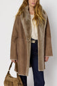 Woman with long wavy hair wearing a tan manteau peau lainée shearling coat over a lace top and dark jeans, holding a tan handbag. The coat features a wide fur collar and a single button.