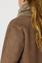 A person with long, wavy blonde hair wearing a brown suede manteau réversible with a high, soft beige collar, shown from the back and side against a plain background.