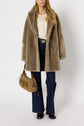 A woman stands wearing a chic manteau col châle in tan faux fur, a cream lace blouse, dark blue flared jeans, and black boots, holding a tan handbag.
