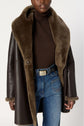 A person wearing a dark brown manteau peau lainée with a large fur collar, a brown turtleneck sweater, blue jeans, and a light-colored belt.