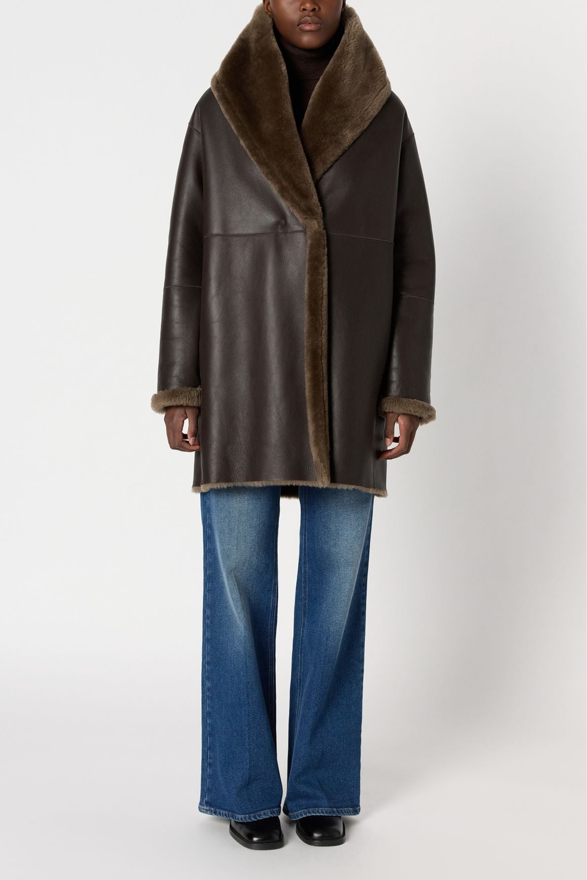 A person wearing a manteau peau lainée—specifically, a dark brown shearling coat with a wide fur collar and cuffs—paired with blue flared jeans and black shoes, stands against a plain light background.