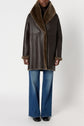 A person wearing a manteau peau lainée—specifically, a dark brown shearling coat with a wide fur collar and cuffs—paired with blue flared jeans and black shoes, stands against a plain light background.