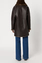 A person wearing a dark brown manteau cuir mouton with a furry collar and cuffs, paired with blue jeans, is shown from the back against a plain white background.
