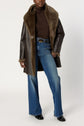Person wearing a brown manteau cuir mouton faux fur-lined leather coat, brown turtleneck sweater, blue high-waisted flared jeans, and black shoes, standing against a plain white background.