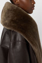 A person wearing a brown manteau cuir mouton with a large, soft, furry collar is shown from the side against a plain background.