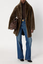 A person wearing a manteau cuir mouton—a chic brown faux fur coat—paired with a brown turtleneck, flared blue jeans, and black shoes holds a brown leather bag against a plain background.
