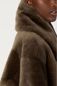 A close-up of a person wearing a plush, brown manteau peau lainée faux fur coat with a large collar. Only part of the person’s face and shoulder are visible against a light background.