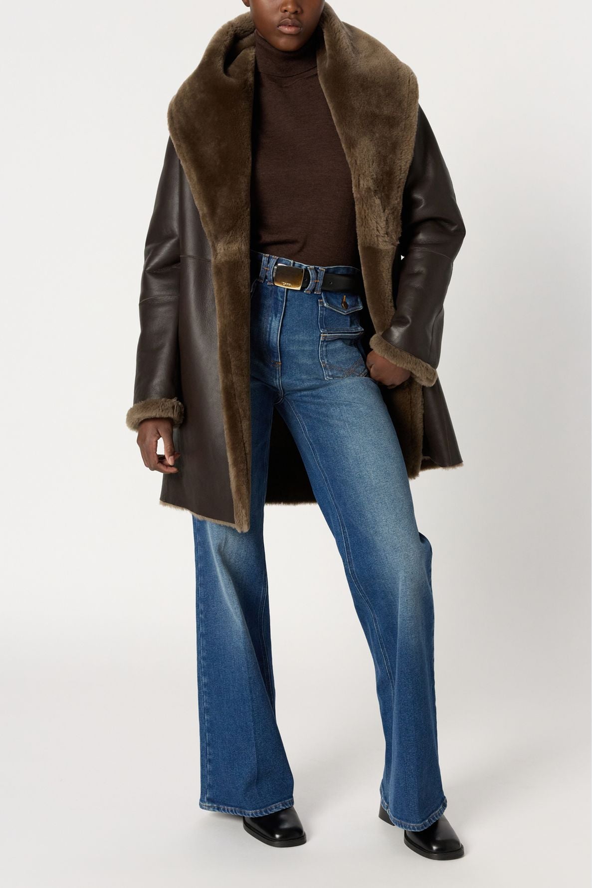 Person wearing a brown shearling coat (manteau cuir mouton), brown turtleneck, blue flared jeans, and black shoes, standing against a plain light background. The manteau cuir mouton features a large fur collar and cuffs.