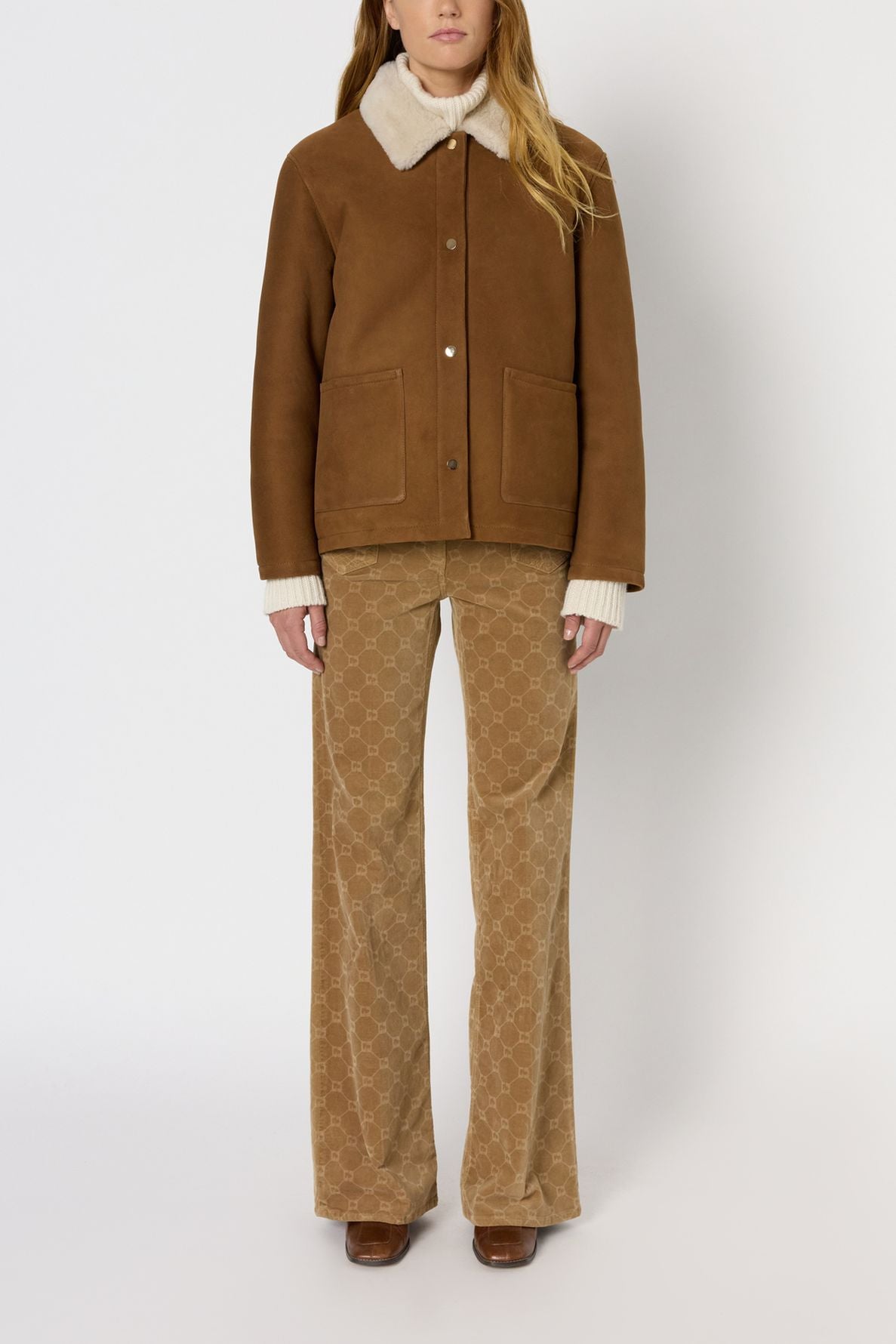 A person wearing a brown shearling jacket reminiscent of a veste cuir femme, beige patterned flared pants, brown shoes, and a cream-colored sweater stands against a plain white background.