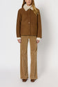 A person wearing a brown shearling jacket reminiscent of a veste cuir femme, beige patterned flared pants, brown shoes, and a cream-colored sweater stands against a plain white background.