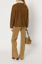 A person stands facing away, wearing a veste camel shearling jacket, beige patterned pants, and brown heeled shoes, holding a tan handbag.