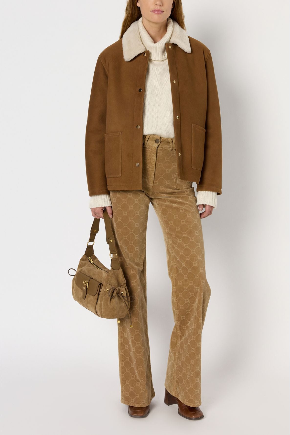 A woman wears a tan veste camel shearling jacket, cream sweater, patterned tan flared pants, brown boots, and carries a tan suede handbag. She stands against a plain white background.