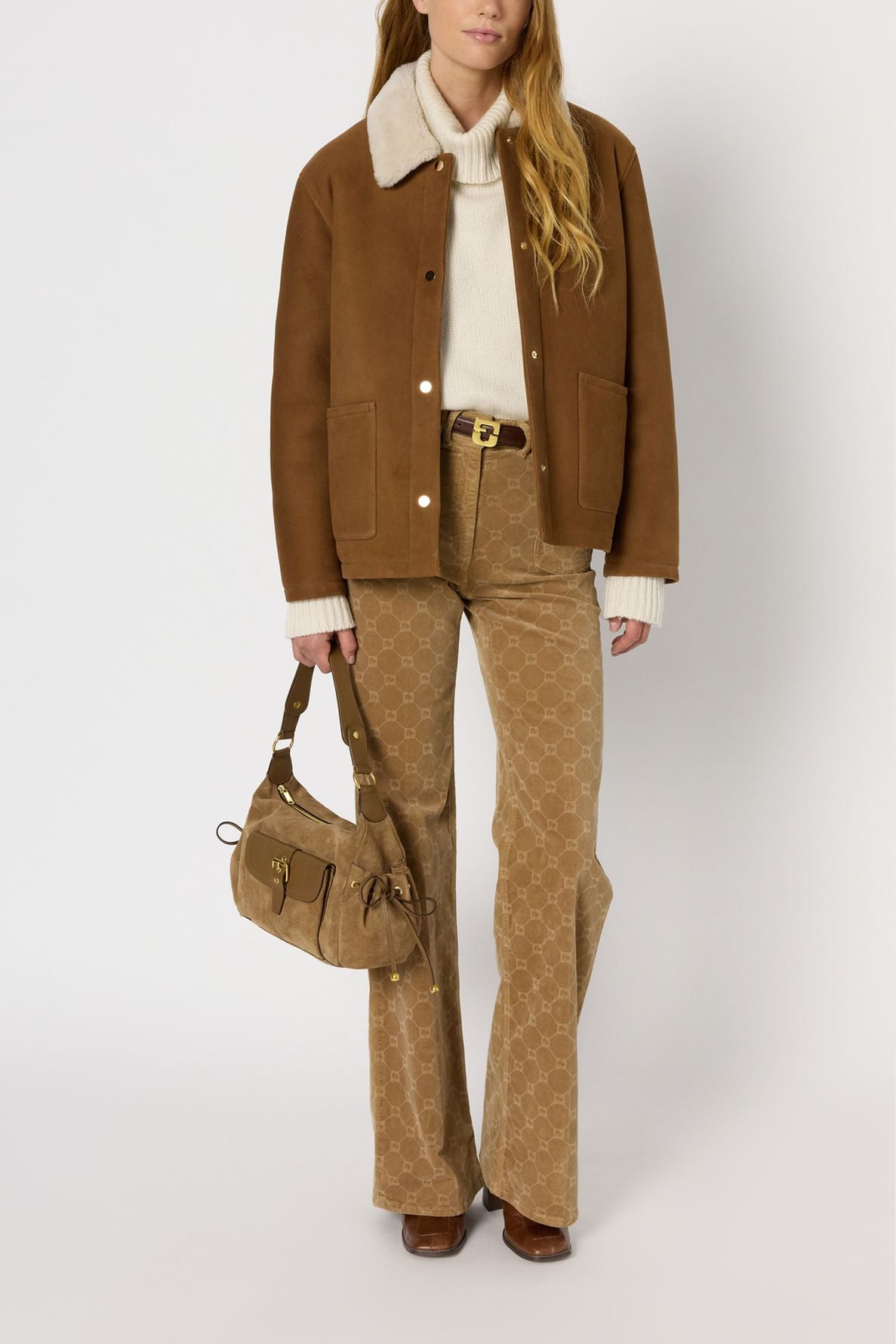 A woman wearing a veste camel with a white collar, cream sweater, tan patterned pants, brown boots, and holding a tan handbag stands against a plain white background.