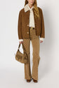 A woman wearing a veste camel with a white collar, cream sweater, tan patterned pants, brown boots, and holding a tan handbag stands against a plain white background.