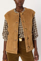 A person wearing a tan veste sans manche buttoned vest over a plaid shirt and brown corduroy pants, holding a bag. The vest, possibly crafted from soft cuir d’agneau, features large buttons and front pockets. The background is plain white.