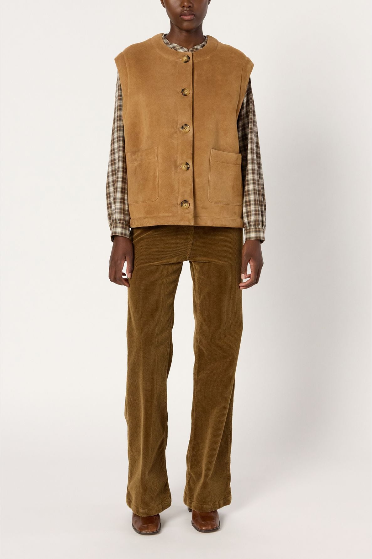 Person wearing a tan veste sans manche in soft cuir d’agneau over a brown plaid shirt, with matching tan corduroy pants and brown shoes, standing against a plain white background.