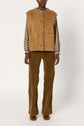 Person wearing a tan veste sans manche in soft cuir d’agneau over a brown plaid shirt, with matching tan corduroy pants and brown shoes, standing against a plain white background.