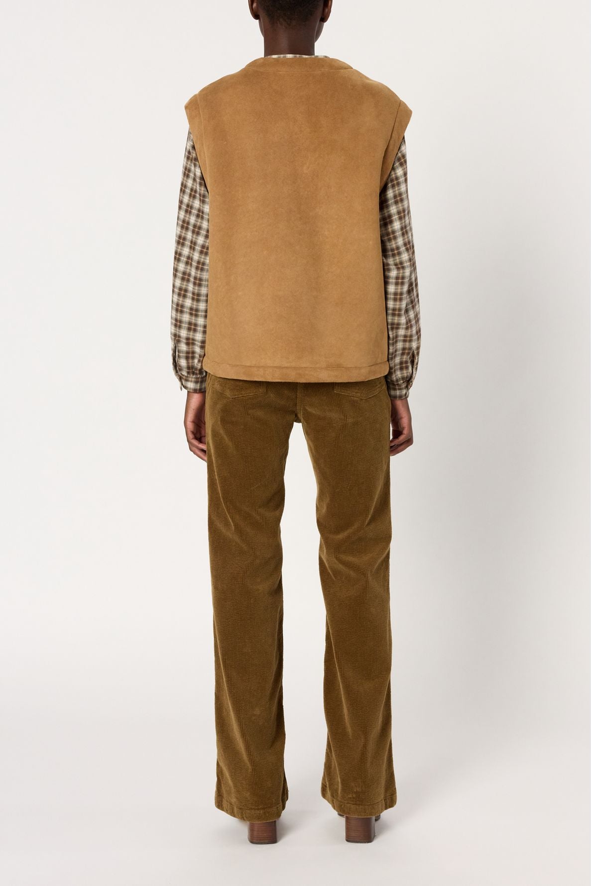 Person standing facing away, wearing a veste sans manche in tan suede cuir d’agneau over a brown plaid shirt and wide-legged brown corduroy pants, against a plain white background.