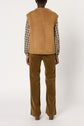 Person standing facing away, wearing a veste sans manche in tan suede cuir d’agneau over a brown plaid shirt and wide-legged brown corduroy pants, against a plain white background.