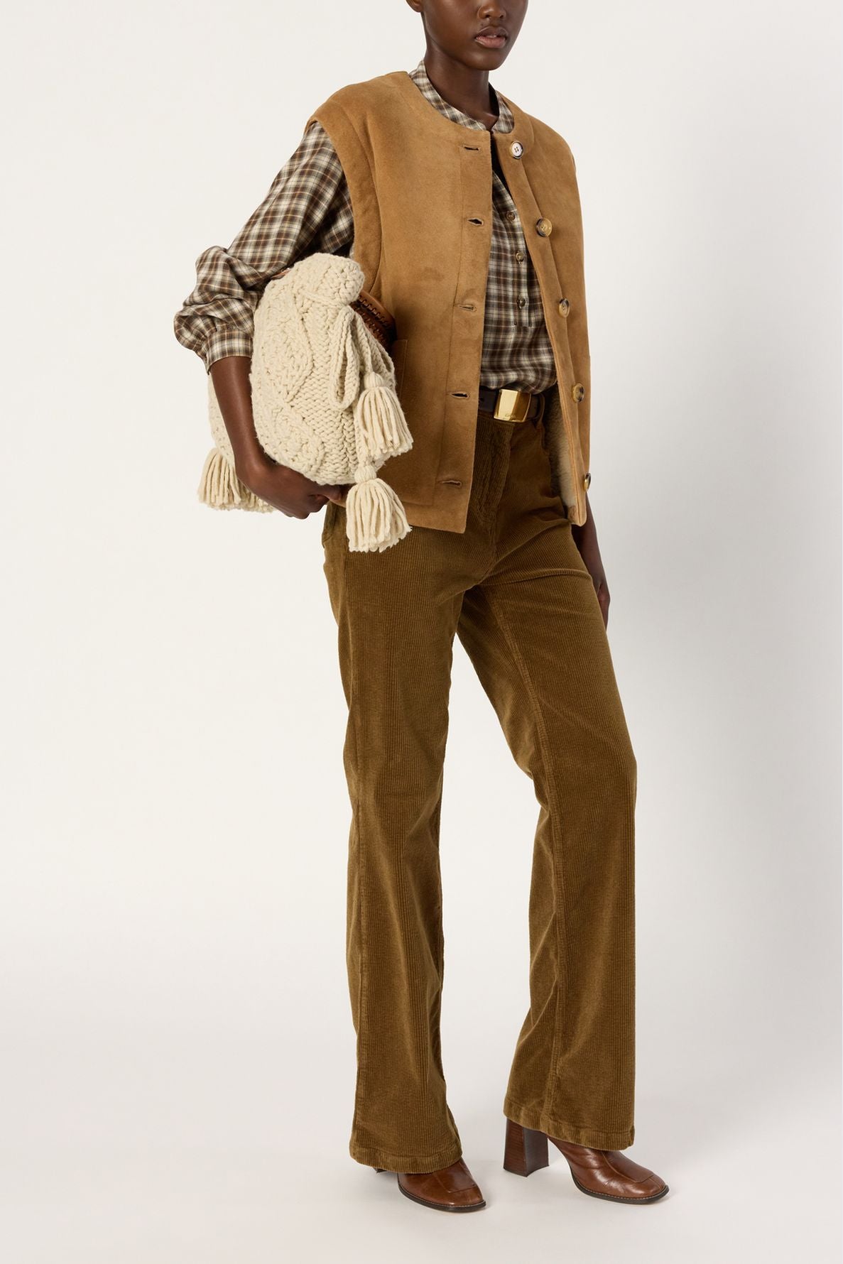 Person wearing a tan veste sans manche, brown plaid shirt, and brown corduroy pants with brown shoes, holding a cream-colored knit bag with tassels. The background is plain white.