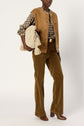 Person wearing a tan veste sans manche, brown plaid shirt, and brown corduroy pants with brown shoes, holding a cream-colored knit bag with tassels. The background is plain white.
