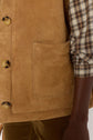 Close-up of a person wearing a tan suede veste sans manche with a large front pocket, over a brown, white, and gray plaid shirt. Part of their arm and brown corduroy pants are also visible.