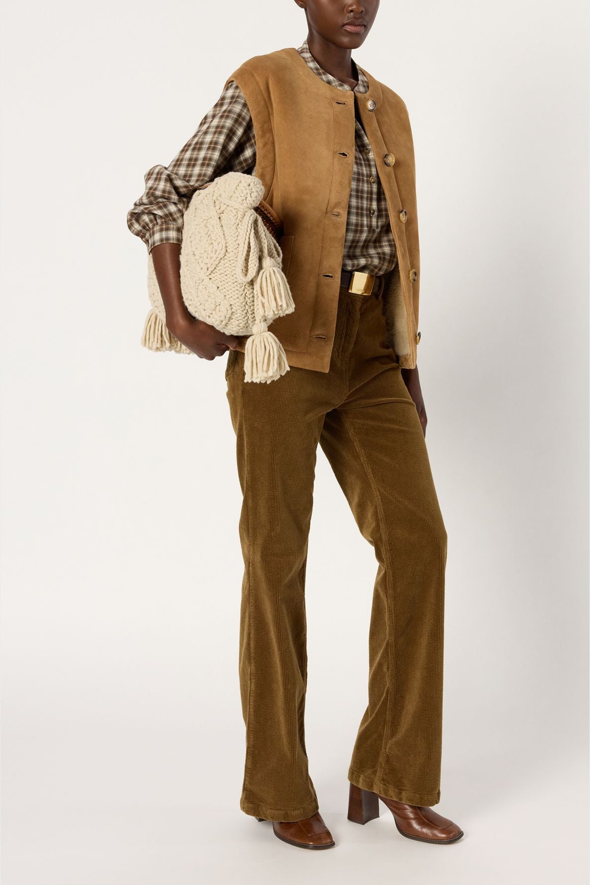 A person wearing a brown suede veste sans manche, plaid shirt, corduroy pants, and brown boots holds a large cream knitted bag with tassels. Background is plain white.