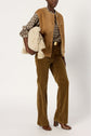 A person wearing a brown suede veste sans manche, plaid shirt, corduroy pants, and brown boots holds a large cream knitted bag with tassels. Background is plain white.