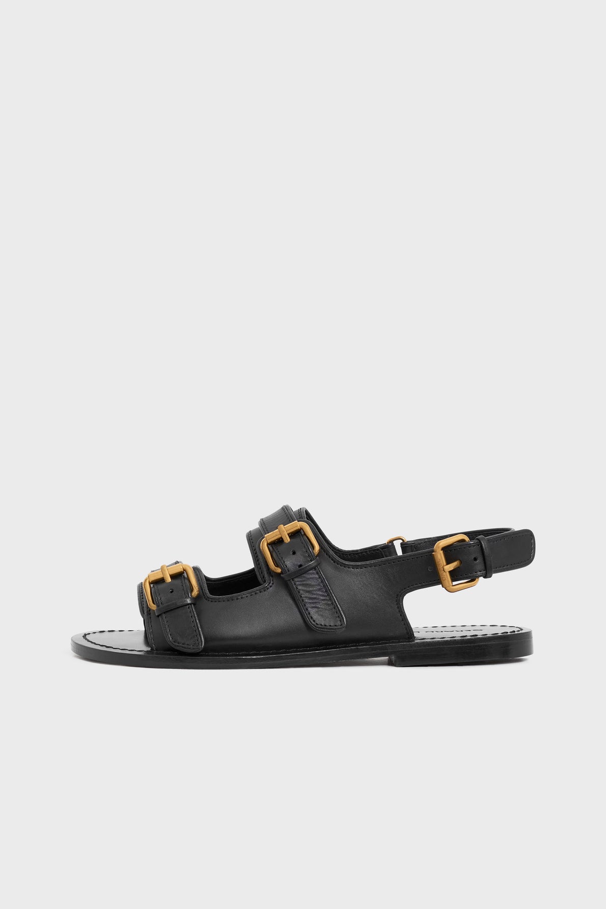 Black sandales cuir with two gold buckles on the front straps and one on the ankle strap, featuring a flat sole and an open heel. These elegant sandales à boucles are displayed on a plain light gray background.