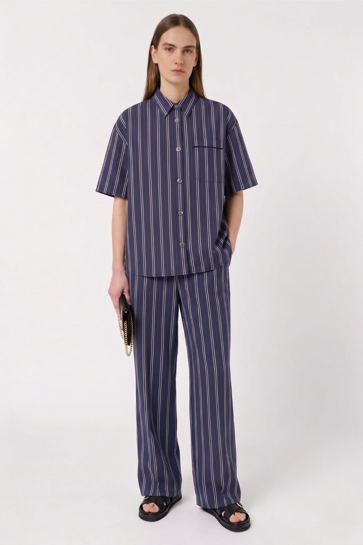 Person with long hair wears a matching navy blue pinstripe short-sleeve shirt—chemise bleu nuit à rayures femme—and wide-leg pants, holds a clutch bag, and stands against a plain light background.