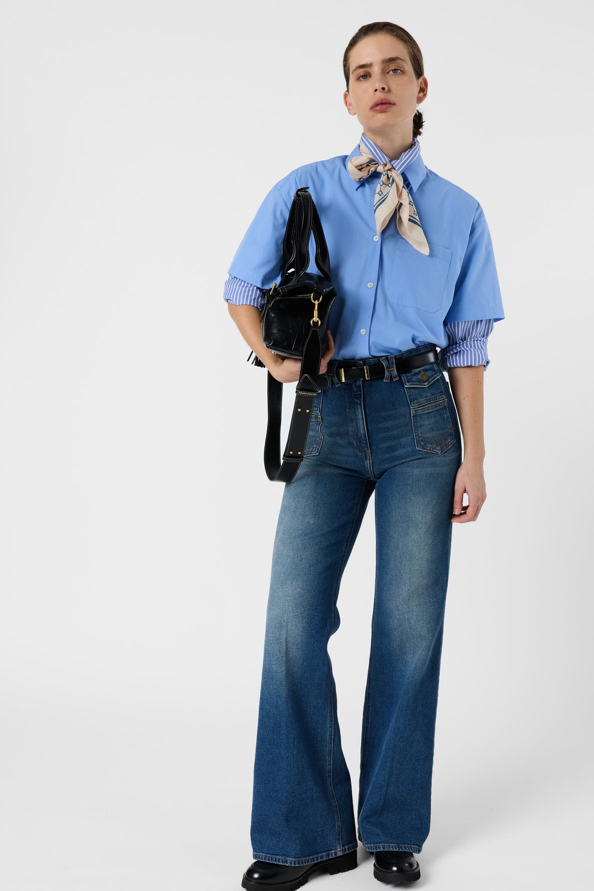 A person wearing a chemise ample bleue, flared blue jeans, black shoes, a patterned neck scarf, and holding a black shoulder bag stands against a plain white background.