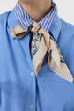 A person wearing a chemise ample bleue button-up shirt with a blue and white striped collar, accessorized with a beige and navy patterned silk scarf tied in a knot around the neck.