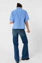 Person standing with their back to the camera, wearing a chemise ample bleue à manches courtes superposée sur un sous-pull, accompagnée d’un jean bleu et de chaussures noires, le tout posé sur un fond blanc épuré.