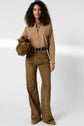 A woman with curly hair wears a chemise popeline camel, brown bell-bottom pants, and tan shoes. She holds a brown bag in her left hand and stands facing forward against a plain background.